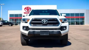 Professional photograph of a 2025 Toyota Tacoma truck parked at a modern dealership lot, showing the vehicle's front three-quarter angle with clear detail of the grille, headlights, and truck bed, bright daylight with blue sky background