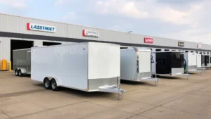 Professional enclosed cargo trailer parked in commercial logistics yard with business signage and modern warehouse facility in background, showing different trailer sizes and configurations