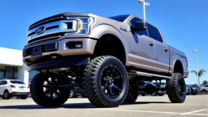Professional photograph of a lifted Ford F-150 truck elevated on suspension system, parked in outdoor dealership lot with clear blue sky, showcasing the lift height and wheel upgrades from side angle