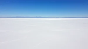 Aerial view of vast white salt flat extending to horizon under clear blue sky with distant mountains, photorealistic landscape photography, no text or signage visible