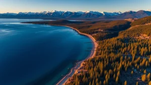 Aerial photograph of Lake Tahoe shoreline with snow-capped mountains, clear blue water, and mixed forest landscape during golden hour, photorealistic professional real estate photography style, no text or signage visible