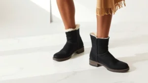 Professional female model wearing classic black UGG boots paired with neutral winter outfit, standing confidently on white marble floor, natural studio lighting, high-quality product photography style